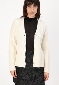 Cardigan cremoso a coste con scollo a V profondo, grandi bottoni e spalle scese, abbinato a un top nero trasparente e una gonna fantasia.