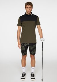 Olivi vihreä polo-paita mustalla yläpaneelilla, yhdistettynä mustiin kamouflageshortsit. Malli pitää golfmailaa ja käyttää valkoisia sukkia mustilla raidoilla.