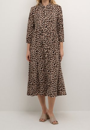 Frau trägt ein langärmeliges Midi-Kleid mit beigen und schwarzen Leopardenmuster und beige offene Blockabsatz-Sandalen, steht vor einem einfarbigen Hintergrund.