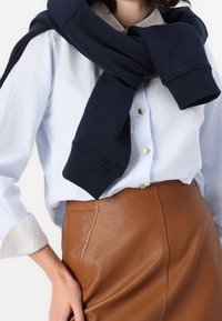 Donna con camicia azzurra chiara a bottoni infilata in una gonna di pelle marrone, maglione blu navy appoggiato sulle spalle, mano appoggiata sui fianchi.