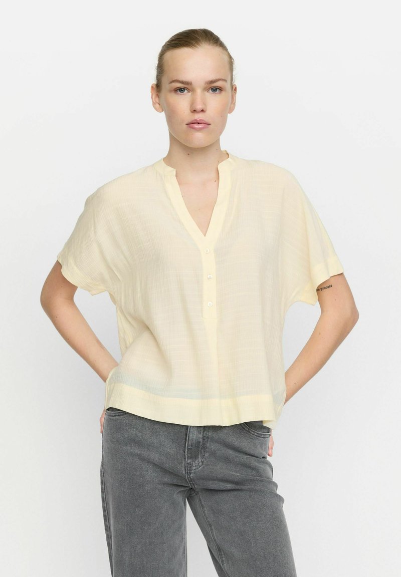 Lichtgele blouse met korte mouwen en een V-halslijn, voorzien van een knopenlijst. Getextureerde stof met een losse pasvorm, weergegeven op een neutrale achtergrond.