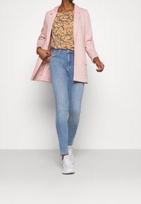 Eine Frau trägt ein hellrosa Blazer, eine blumige gelbe Bluse, hellblaue Skinny-Jeans und weiße Sneaker und geht vorwärts vor einem plain weißen Hintergrund.