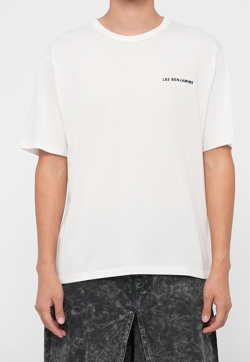 Personne portant un t-shirt blanc uni à manches courtes avec le texte "LES BENJAMINS" sur la poitrine gauche et un pantalon noir délavé à l'acide.