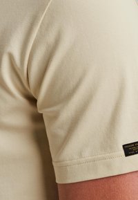 Camiseta de algodón beige con cuello redondo, mangas cortas, textura suave y una pequeña etiqueta negra en la costura lateral inferior.