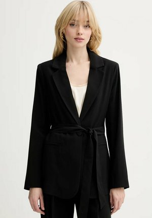 Donna bionda che indossa un blazer nero con cintura sopra una maglietta bianca, in piedi davanti a uno sfondo chiaro e uniforme, rivolta in avanti con espressione neutra.