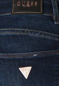 Jeans de mezclilla azul oscuro con costuras en naranja contrastante, un parche de cuero con "GUESS" en relieve y un detalle de logo metálico triangular.