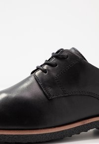 Chaussure en cuir noir avec une finition lisse, bout rond et lacets. Présente des accents cousus et une semelle en caoutchouc texturée avec un bord marron clair.