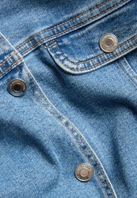 Lähellä oleva kuva sinisestä farkkukankaasta, jossa on oranssia tikkausta ja kolme metallista nappia, joissa lukee "DENIM."