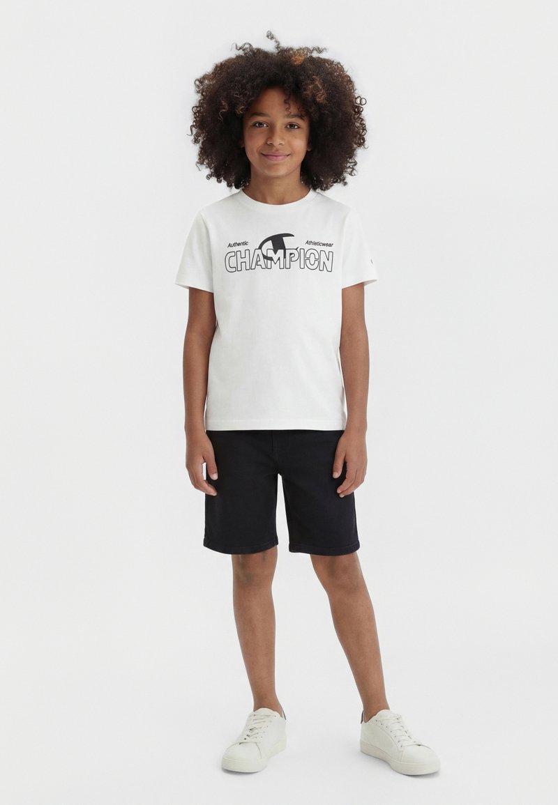 Enfant aux cheveux bouclés portant un t-shirt blanc Champion, un short noir et des baskets blanches, debout devant un fond uni clair.