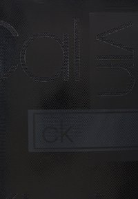 Surface texturée sombre avec des lettres en relief formant le mot "Calvin" et un petit rectangle contenant "ck" au centre.