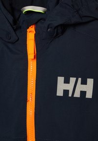 Marineblauwe jas met een opvallend zilveren logo, voorzien van een oranje ritssluiting en details. Gemaakt van een gladde, duurzame stof. Met capuchon.