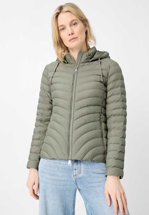 Frau trägt eine hell olivgrüne gesteppte Kapuzenjacke mit Frontreißverschluss und hellblaue Jeans, stehend vor weißem Hintergrund.