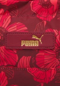 Tissu à motif floral rouge avec de grandes fleurs et feuilles, mettant en avant un logo "PUMA" doré sur une étiquette en simili cuir bordeaux.