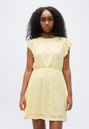 Lokkis juustega naine, kes kannab lühikest, helekollast õhulist kleiti võrkdetailidega ja lendlevate varrukatega, seisab tavalise heleda tausta ees.