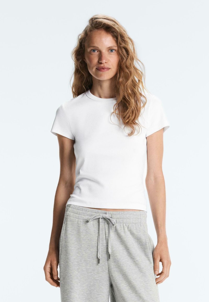Femme aux cheveux longs et ondulés portant un t-shirt blanc uni à manches courtes et un pantalon gris clair à cordon, de face avec une expression neutre.