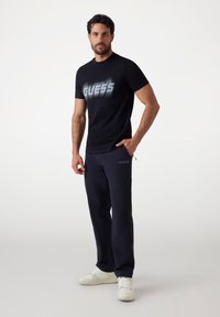 Zwarte korte mouwen t-shirt met het "GUESS" logo in een gradiënt ontwerp, gecombineerd met donkere broek en witte sneakers. Eenvoudige, atletische pasvorm.