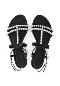 Schwarze flache Sandalen mit mit Perlen verzierten Riemen, Knöchelriemen und kleinen schwarzen Schleifen oben, von oben auf einem weißen Hintergrund gezeigt.