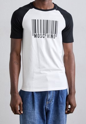 Mężczyzna w białej koszulce z krótkim rękawem, czarnymi rękawami raglan i grafiką kodu kreskowego z tekstem "Moschino" na piersi.