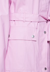 Rosa, wasserdichte Jacke mit tailliertem Gürtel, großer aufgesetzter Tasche, Druckknöpfen und glatter, reflektierender Oberfläche.