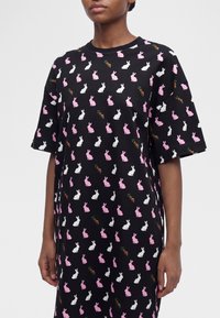 Μαύρο oversized φόρεμα πουκάμισο με κοντά μανίκια και σχέδια ροζ και λευκού λαγού και καρότου, που φοριέται από ένα άτομο με τα χέρια χαλαρά στο πλάι.