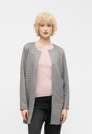 Femme aux cheveux blonds bouclés portant une veste matelassée grise ouverte sur un t-shirt rose clair et un pantalon noir sur un fond blanc.