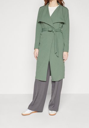 Vrouw draagt een lange groene jas met riem, grijze wijde broek, wit shirt en witte sneakers tegen een eenvoudige lichte achtergrond.