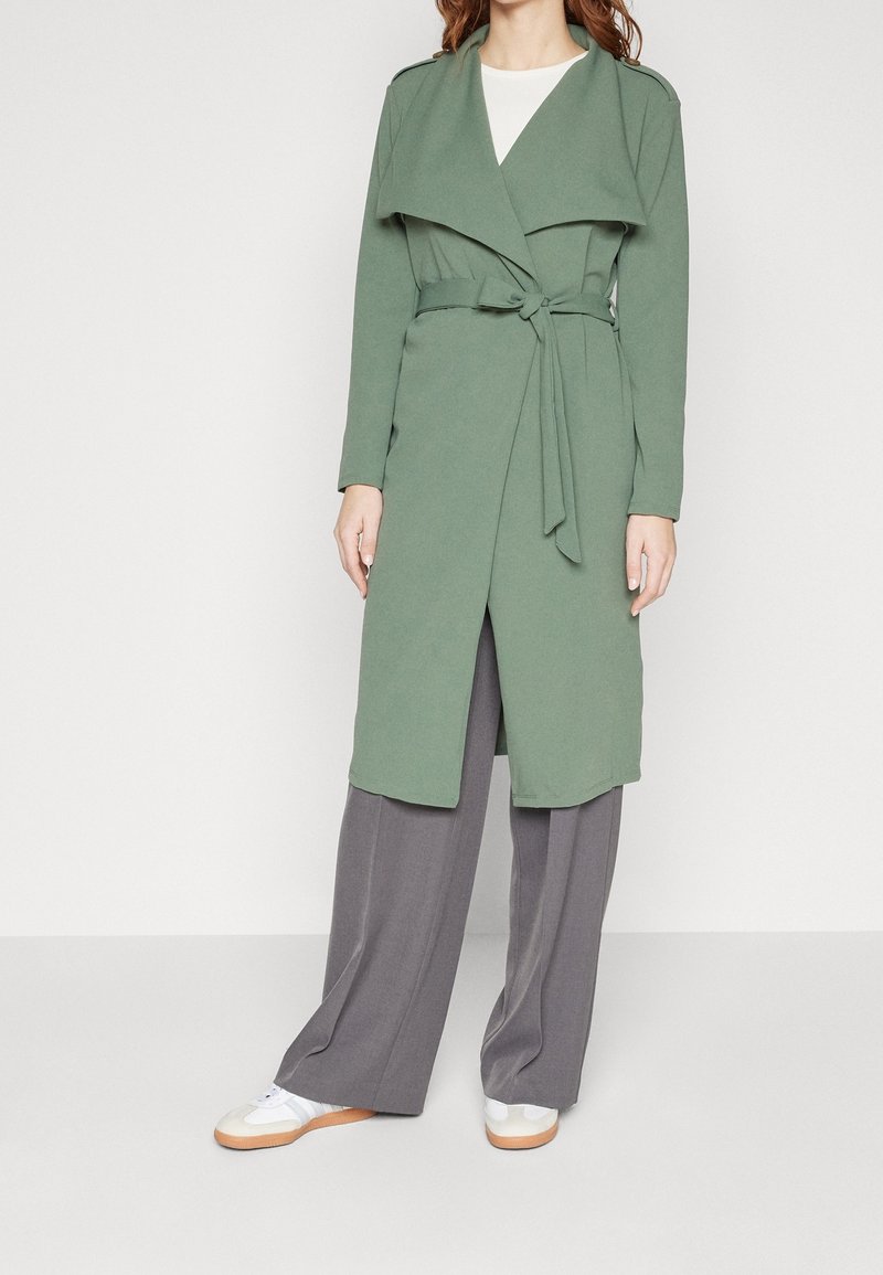 Femme portant un long manteau vert ceinturé, un pantalon gris à jambes larges, une chemise blanche et des baskets blanches sur un fond clair uni.