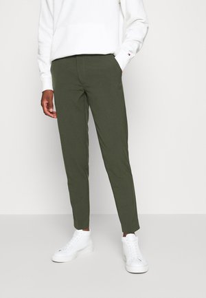 Person trägt olivgrüne Slim-Fit-Hose, weiße High-Top-Sneaker und ein weißes Langarmshirt mit einer Hand in der Tasche.