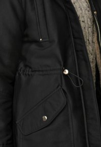 Veste noire en tissu doux avec une fermeture éclair, une poche latérale et un cordon de serrage réglable au niveau de la taille. Comprend des accents métalliques et des coutures.