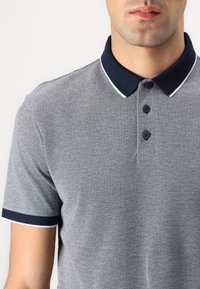 Siva polo majica s mornarsko plavim ovratnikom i detaljima, s tri crna dugmeta i teksturiranom tkaninom. Prikaz izbliza.