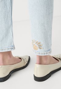 Lyseblå denimjeans med dekorativ brunt broderi ved kanten, paret med beige flate sko med svarte såler og dekorative detaljer.