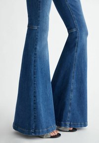 Jeans in denim svasato con lavaggio blu scuro, caratterizzati da vita alta, gamba larga e cuciture a contrasto sulle cuciture. Tessuto con trama visibile del tessuto.
