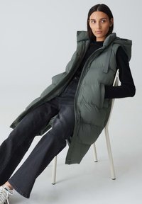 Langer, kapuzenloser Steppwesten in Dunkelgrün, mit gestepptem Muster und Knopfverschluss auf der Vorderseite. Trägt man über einem schwarzen Oberteil und kombiniert mit dunklen Schlagjeans.