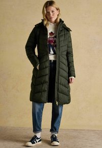 Tmavozelený prešívaný dlhý kabát s kapucňou, predný zips, bočné vrecká, skombinovaný so svetlomodrými džínsami a námorníckymi teniskami. Pod ním je textúrovaný pletený sveter.