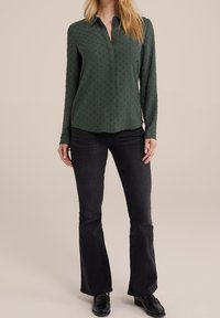 Mörkgrön skjorta med knappar och texturmönster, långa ärmar och en rundad fåll, i kombination med svarta utsvängda jeans och svarta skor.