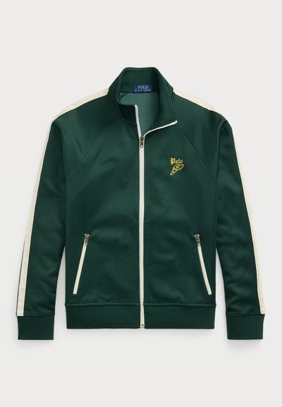 Grønn zip-up jakke laget av glatt stoff, med hvite side-striper og et gull-logo på venstre bryst. Inkluderer to glidelåslommer.