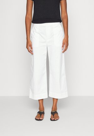 Femme portant un haut noir sans manches, un pantalon blanc à jambes larges et raccourci avec poches avant, et des sandales noires à enfiler avec des lettrages dorés.