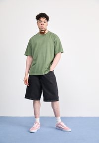 Camisa verde de manga corta, pantalones cortos de denim negros y zapatillas rosas con suelas blancas. Atuendo casual de una persona con cabello rizado. Fondo claro.