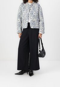 Veste matelassée avec motif paisley bleu marine sur fond blanc ; associée à un pantalon noir à jambes larges et des talons pointus noirs ; tenant un petit sac noir.