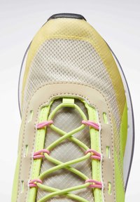 Chaussure de course légère avec une tige en mesh beige ornée d'accents jaunes, des lacets verts et roses, et un design texturé pour une meilleure adhérence.