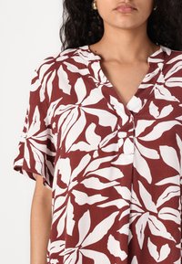 Chemise rouge foncé à manches courtes avec un motif floral blanc, dotée d'un col en V et d'un détail de bouton au col. Texture de tissu légère.