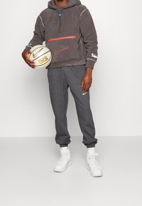 Felpa grigia in pile con tasca con zip e accenti arancioni, abbinata a pantaloni jogger grigi e sneakers bianche alte. Tiene un pallone da basket.