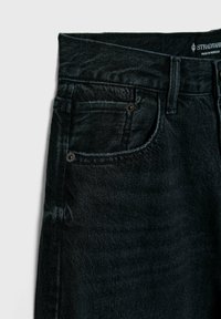 Jean en denim noir en gros plan montrant la poche avant, les passants de ceinture, les détails de coutures, et une partie de la ceinture avec l'étiquette de la marque.