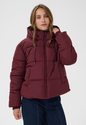 Femme portant une veste matelassée bordeaux avec une capuche, debout avec une main près du visage et un jean bleu foncé.