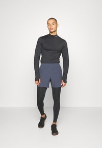 Svart langermet topp, marineblå shorts over svarte leggings, og svarte joggesko. Designet er tettsittende med tommelhull og elastisk midje.