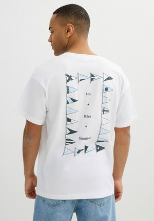 Hombre con camiseta blanca con banderas náuticas en azul claro y negro dispuestas en un rectángulo y texto "Les Belles Bannières" en la espalda.