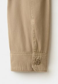 Manche longue beige avec un poignet à bouton unique et une couture détaillée sur tissu en coton.