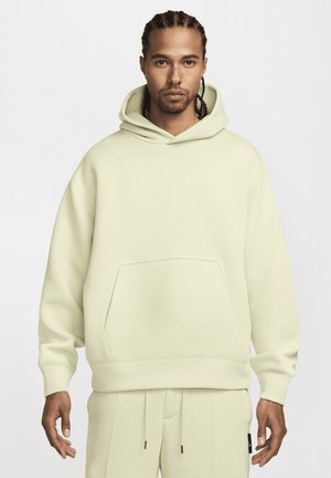 Kottide punutud juustega mees, seljas heledabež või oversize kapuutsiga sweatshirt ja sobivad püksid, seismas ühetaolise valge tausta ees.