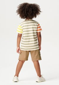 Maglietta a strisce in beige, verde oliva e maniche brillanti; abbinata a pantaloni corti khaki. Il tessuto è cotone con una texture morbida. Stile casual.