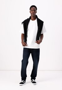 Sort ærmeløs top draperet over en hvid t-shirt, parret med mørkeblå jeans og sorte sneakers med hvide detaljer. Minimalistisk design.